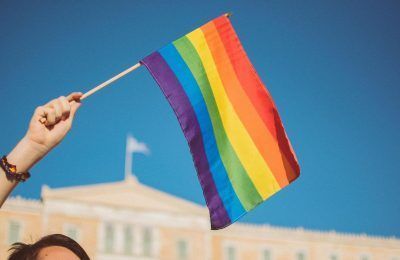 A person holding the iconic rainbow-coloured LGBTQ+ flag with the Greek Parliament building in Syntagma, Athens, in the background. Photo by Stavrialena Gontzou on Unsplash.
